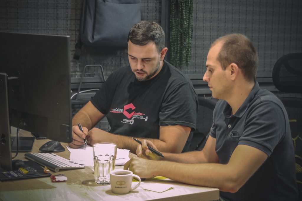 Image shows two man sitting at the desk, looking at papers and working on something in the office